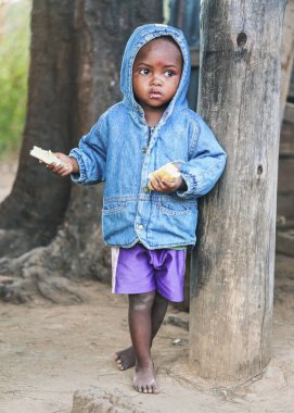 Ranohira, Madagaskar - 29 Nisan 2019: Mavi kot ceketli ve şortlu, tahta sütunda duran, ekmek yiyen, Malagasy 'li tanınmamış genç - bu ülkedeki insanlar fakir ama neşelidir.
