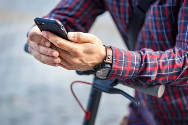 Giovane uomo che indossa una camicia appoggiata al manubrio dello scooter elettrico, tenendo in mano lo smartphone mobile, dettaglio primo piano — Foto Stock
