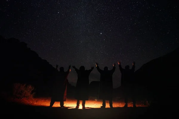 Four persons wearing long warm coats standing in night desert, hands raised to sky with stars, light on the ground, view from behind only silhouettes visible