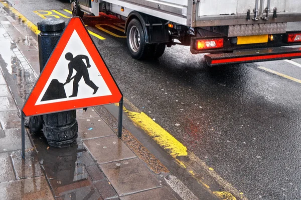 Warning roadworks sign at wet road next to pavement, van moving in background over Bus Stop writing on road