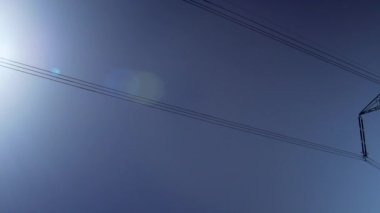 Açık mavi gökyüzüne karşı elektrik direği ve kablolarına bakıyorum. Güneş döndükçe lensler parlıyor.