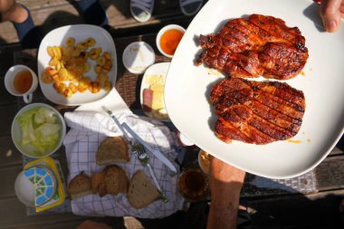 İki taze ızgara domuz filetosu, beyaz tabakta. Bahçe masasının üstünde, ızgaradan daha fazla yiyecek var..