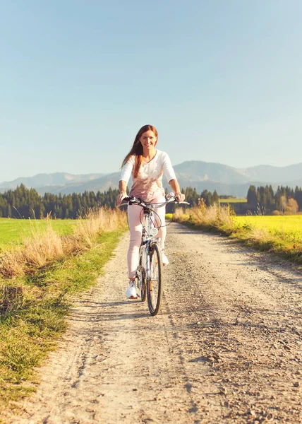 Young woman in summer light clothes rides bicycle on dusty road towards camera, afternoon sun shines to fields and forest in background
