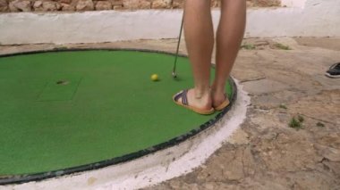 Genç adam mini golf oynuyor, ayakları üzerinde, sandaletlerle, sarı topuyla üç kez deliği ıskalıyor.