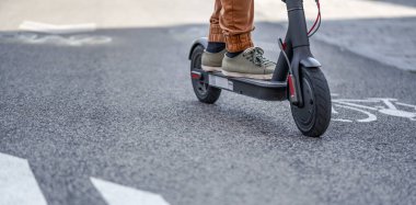 Ayaklarında elektrikli scooter ile asfalt yolda gezen sıradan pantolonlu genç bir adam.