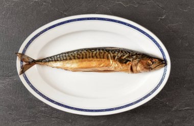 Beyaz oval tabakta tütsülenmiş uskumru balığı, altında masa gibi gri siyah bir taş. Yukarıdan görüntüle