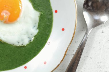Güneşli tarafı beyaz tabakta ıspanakta pişmiş yumurta, yanında küçük kırmızı noktalar, yanında çelik kaşık, yakın çekim fotoğrafı.