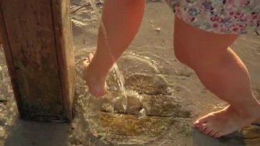 Kumu kesen genç bir kadın kumlu zeminin yakınındaki kumsal duşunun altında bacaklarını oluşturur. Sabah güneşi arka planda parlar. Suyun detayları ayakların üzerine düşer.