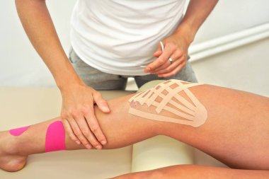 Genç bayan fizyoterapist hastasının dizine kineziyoloji bandı uyguluyor.