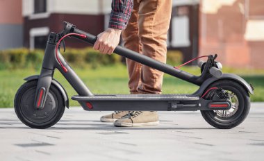 Beton kaldırımda katlanmış elektrikli scooter ön taraftaki bükülmüş barın arkasında duran genç adam, yakın çekim detayı.