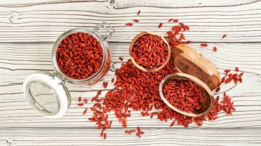 Kurutulmuş goji namı diğer. Kurtüzümü tohumları cam şişenin içinde, bazıları tahta kaselerde ve beyaz tahta masa üzerine dökülmüş, yukarıdan manzara