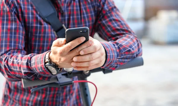 Giovane uomo che indossa una camicia appoggiata al manubrio dello scooter elettrico, tenendo in mano lo smartphone mobile, dettaglio primo piano — Foto Stock