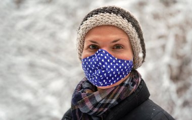 Kışlık ceketli, kalın şapkalı, pamuk virüsü maskeli, küçük kalpli, karla kaplı arka planda kalan ağaçlar, yakın plan detayları.