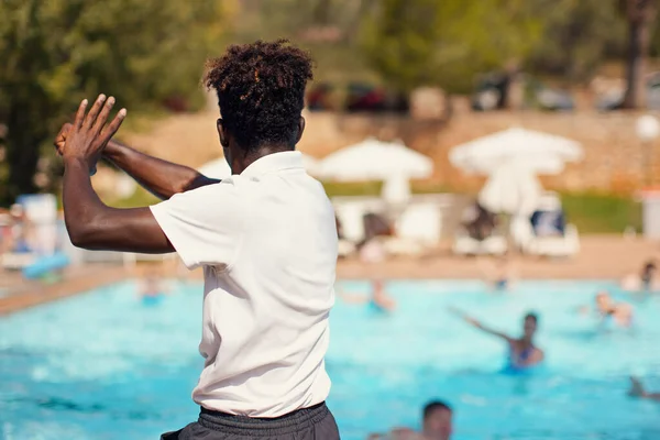 Black athletic hotel resort animator exercising in front of pool, view form behind blurred people background - water aerobic activity