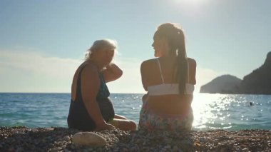 Gri saçlı yaşlı bir kadın güneşli çakıl taşlı plajda oturuyor, genç olanla konuşuyor - kızı, arkadan görüyor.