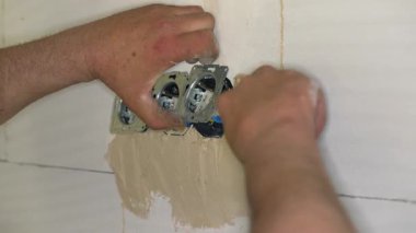 Erkek inşaat işçisi, çıplak duvara havan topu macunuyla elektrik prizleri yerleştiriyor. Delikleri sadece küçük kepçelerle dolduruyor.