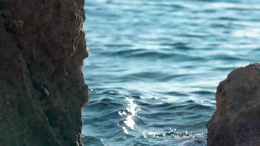 Sahilde oldukça sakin, açık yeşil bir deniz. Büyük kayalar, kıyıdaki pürüzlü dokuya yakın çekim detayları, bulanık su arka planı.