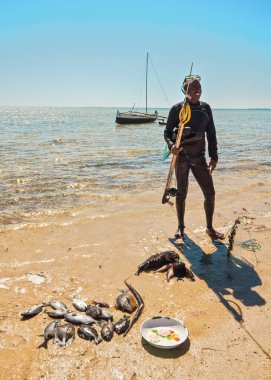 Anakao, Madagascar - May 03, 2019: Local Malagasy fisherman in neoprene wetsuit, with snorkel and simple harpoon, standing next to pile of fresh caught fish at the beach on a sunny day