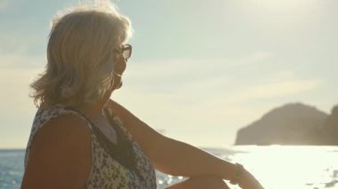 Gözlüklü, sakin deniz kenarında dinlenen yaşlı bir kadın, arka planda güneş ışığı, yakın çekim detayı.
