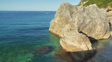 Kıyıda büyük kayalarla sakin, berrak yeşil deniz, güneş küçük dalgalarda parlıyor, kamera yan yana, Yunanistan 'ın Liapades kentindeki pek çok koydan birinde tipik manzara