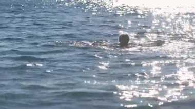 Gri saçlı yaşlı bir kadın, kıyıya yakın denizde yüzüyor, öğleden sonra güneşi arkadan su yüzeyine yansıyor.