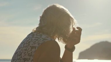 Sakin deniz kıyısında dinlenen yaşlı bir kadın. Gri saçlarını düzeltiyor. Arka planda güneş ışığı var.