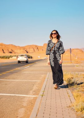 Uzun siyah elbiseli genç bir kadın yol kenarında yürüyor. Arka planda AlUla 'nın tipik çöl kayalık manzarası var.