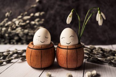 white eggs with a cute smile in a small wooden barrel with snowdrops and pussy willow branches with seals on a gray background. Easter card for the holiday
