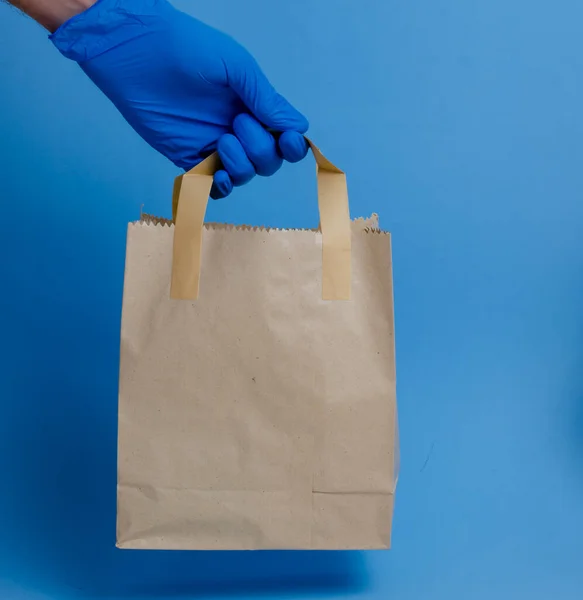brown paper shopping bag on blue background. Brown empty craft lunch bag. Recycle brown paper bag. Hand in glove holding a paper bag.