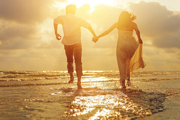 Молодая пара, держась за руки и бегая по пляжу и закату.