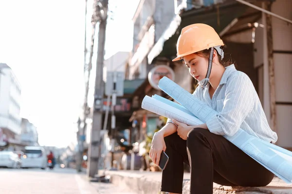 Architecture woman tired and sad and him sitting on the ground and hold blueprints design plan and wearing safety helmet at site construction in hot weather day. Architecture, engineering, business and civil concept.