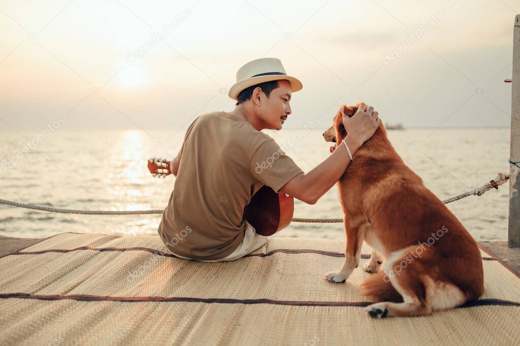 A man wear straw hat and playing guitar music song near the sea sunset and stroking the head dog pet
