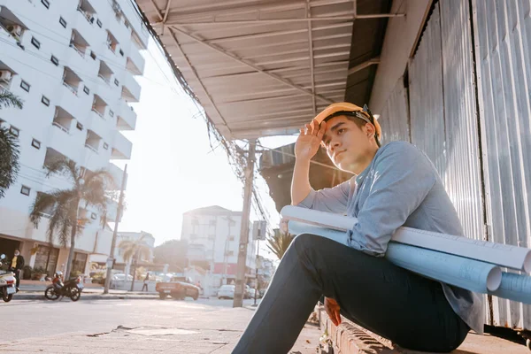 Architecture man wipe the sweat and him sitting on the ground and hold blueprints design plan and wearing safety helmet at site construction in hot weather day. Architecture, engineering, business and civil concept.