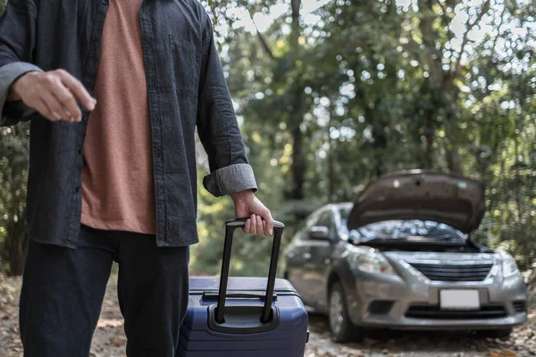 Man' hand drag to travel bag in front of open hood of a broken car on the road in the forest. Car breakdown concept.