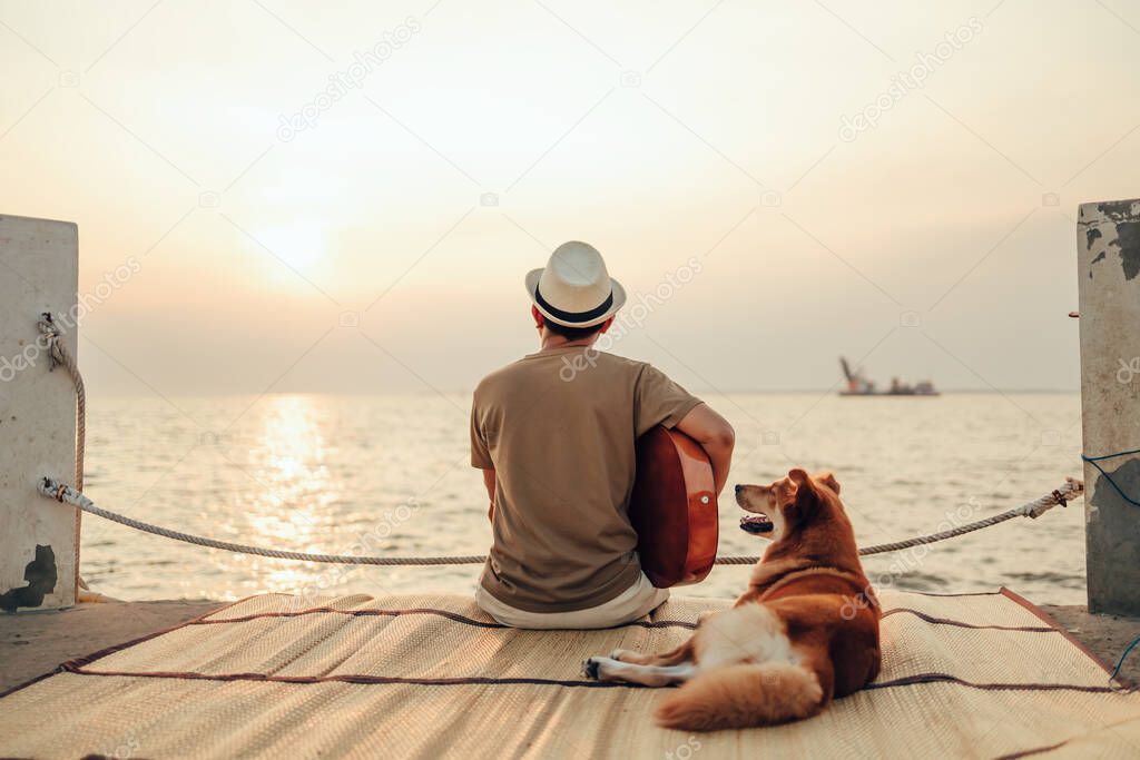 A man wear straw hat and playing guitar music song near the sea sunset with a dog pet
