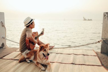 Hasır şapka takan ve deniz kenarında bir köpekle gitar çalan bir adam.