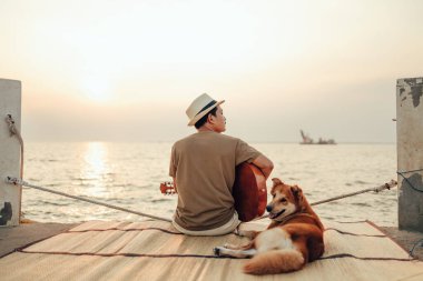Hasır şapka takan ve deniz kenarında bir köpekle gitar çalan bir adam.