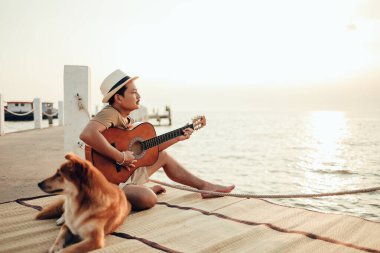 Hasır şapka takan ve deniz kenarında bir köpekle gitar çalan bir adam.