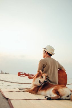 Hasır şapka takan ve deniz kenarında bir köpekle gitar çalan bir adam.