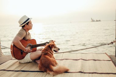 Hasır şapka takan ve deniz kenarında bir köpekle gitar çalan bir adam.