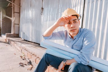 Mimarcı adam terini silerken o da yerde oturuyor ve tasarım planlarını elinde tutuyor ve sıcak hava günlerinde inşaat alanında kaskını takıyor. Mimarlık, mühendislik, iş ve medeni konsept.