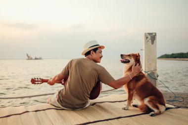 Hasır şapka takan ve deniz kenarında gitar çalan bir adam gün batımına yakın bir yerde evcil hayvanın başını okşuyor.