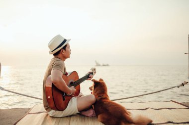 Hasır şapka takan ve deniz kenarında bir köpekle gitar çalan bir adam.