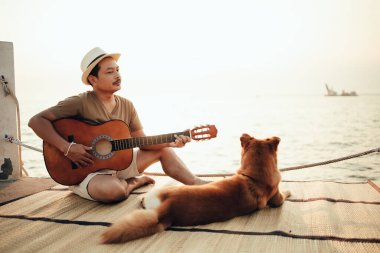 Hasır şapka takan ve deniz kenarında bir köpekle gitar çalan bir adam.
