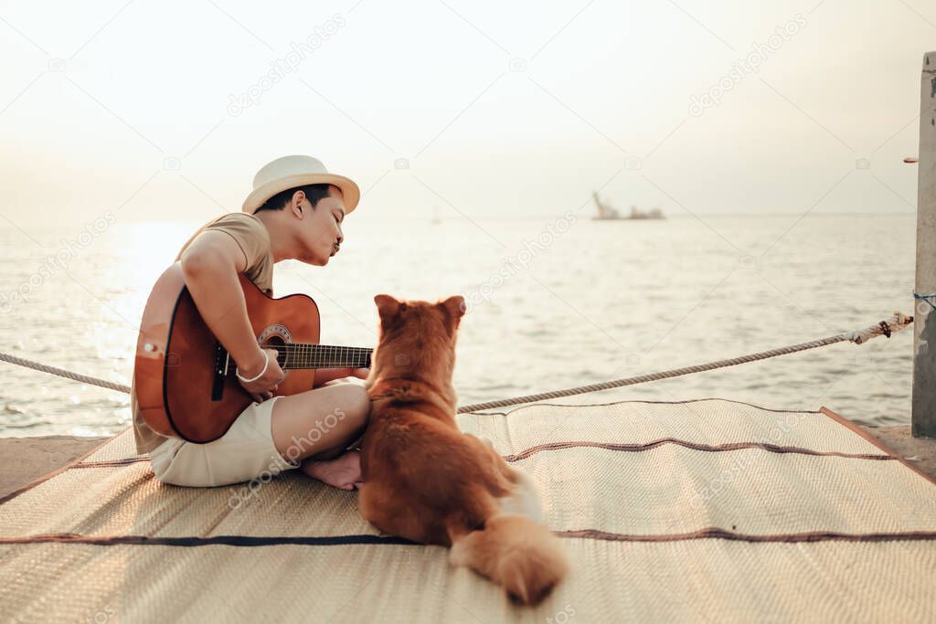 A man wear straw hat and playing guitar music song near the sea sunset and him need kiss with a dog pet