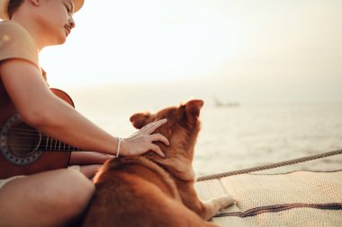 Hasır şapka takan ve deniz kenarında gitar çalan bir adam gün batımına yakın bir yerde evcil hayvanın başını okşuyor.