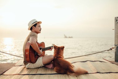 Hasır şapka takan ve deniz kenarında bir köpekle gitar çalan bir adam.