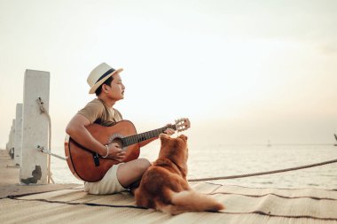 Hasır şapka takan ve deniz kenarında bir köpekle gitar çalan bir adam.