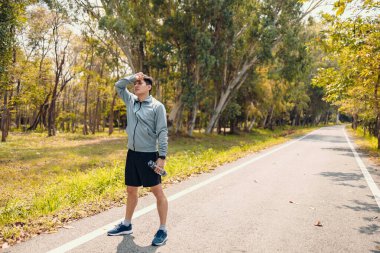 Yorgun bir genç adam terini siliyor. Adam dinleniyor, nefes alıyor ve parkta bir şişe içme suyu tutuyor..