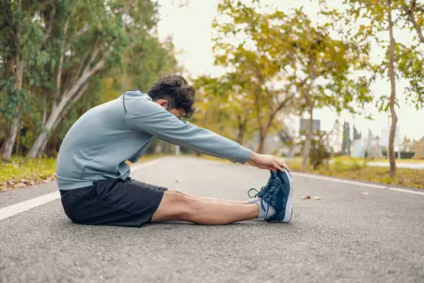 Genç adam koşmadan önce parkta geriniyor. Genç adam parktaki fitness antrenmanından önce antrenman yapıyor. Sağlıklı ve egzersiz yapan genç adam ormanın yanındaki yolda ısınıyor..